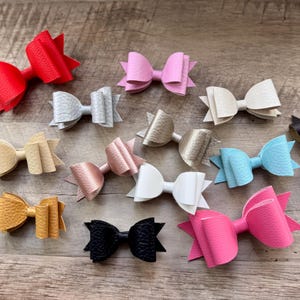 May include: A collection of colorful faux leather bows in various shades, including red, pink, gold, and black. The bows are arranged on a wooden surface, showcasing their textured appearance and layered design. These accessories are ideal for hair styling.