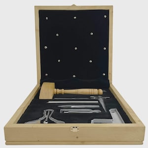 Puede incluir: Caja de madera abierta con interior de terciopelo negro, que contiene un mazo de madera y varias herramientas plateadas. El interior de la caja tiene pequeños tachuelas plateadas. Las herramientas están cuidadosamente dispuestas. La caja es de madera clara.