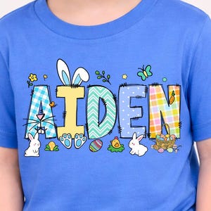 May include: A blue t-shirt with the name "AIDEN" in colorful, patterned letters. The design includes Easter-themed elements like bunnies, eggs, and flowers. The shirt is worn by a young child.