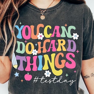May include: A dark gray t-shirt with the text "YOU CAN DO HARD THINGS" in colorful, retro-style lettering. The design includes a pencil, apple, and flowers, with the hashtag #testday.