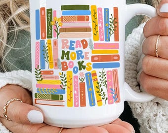 May include: White ceramic mug with a colorful design of stacked books and the words "READ MORE BOOKS" in a playful font. The mug features a variety of book spines in shades of blue, orange, green, and pink, with floral accents.