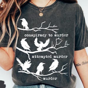 May include: Dark gray t-shirt with a graphic of white ravens on branches, with the words "conspiracy to murder," "attempted murder," and "murder" in white text. The shirt is labeled "pepper" at the bottom.