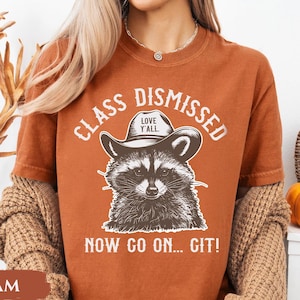 May include: A burnt orange t-shirt with a raccoon wearing a cowboy hat graphic. The text on the shirt reads "CLASS DISMISSED" and "NOW GO ON... GIT!" with "LOVE Y'ALL" on the hat. The shirt is a relaxed fit.