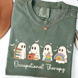 May include: Moss green t-shirt with a crew neck and short sleeves. The shirt features a graphic of four cartoon ghosts with occupational therapy tools and the words "Occupational Therapy" in white script. A gold necklace is draped across the shirt.