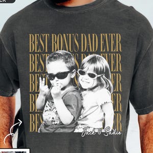 May include: Dark gray t-shirt with a sepia-toned image of two children wearing sunglasses. The text "BEST BONUS DAD EVER" is printed in gold. A smaller photo with the text "CUSTOM PHOTO" is in the lower left.