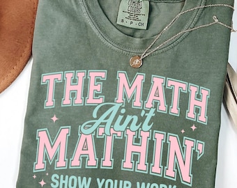 Può includere: T-shirt verde muschio con la scritta "THE MATH Ain't MATHIN' SHOW YOUR WORK" in rosa e turchese. Una collana d'oro con un ciondolo è drappeggiata sulla maglietta. Un cappello marrone e foglie di palma secche sono sullo sfondo.