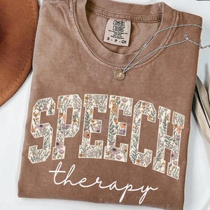 May include: Espresso-colored t-shirt with the words "SPEECH" and "therapy" in white script. The letters of "SPEECH" are filled with a floral pattern. A silver necklace with a pendant is on the shirt. A brown hat is in the background.