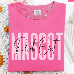 May include: A bright pink t-shirt with the words "MASCOT Pink Out" in a bold, playful font. The shirt has a crew neck and short sleeves. A delicate silver necklace with small charms rests on the shirt. The word "CRUNCHBERRY" is in a pink rectangle in the top right corner.
