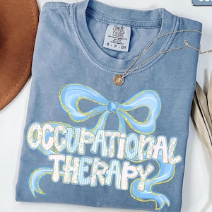 May include: Blue jean Comfort Colors t-shirt with the words "OCCUPATIONAL THERAPY" in a playful font, accented by a light blue bow graphic. A gold necklace with a pendant rests on the shirt. A brown hat is visible in the upper left corner.