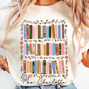 May include: An ivory short-sleeve t-shirt featuring a graphic print of three rows of colorful books arranged vertically, surrounded by a floral border. The text "Mrs. Charlotte" is printed in black cursive script below the book illustration.