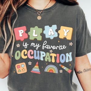 May include: A charcoal gray t-shirt featuring the text "PLAY is my favorite OCCUPATION" in colorful, retro-style lettering. The design includes puzzle piece shapes, a shape sorter, a stacking toy, a rainbow, and a telescope graphic.
