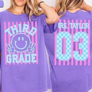 May include: A violet t-shirt featuring a front graphic with pink vertical stripes, a light blue smiley face making peace signs, and the text "THIRD GRADE". The back graphic displays pink vertical stripes, "MRS. TAYLOR", and the number "03" in blue.