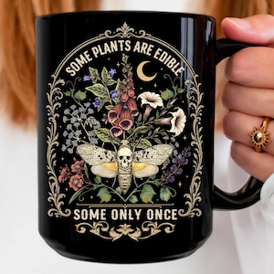 May include: A black ceramic mug featuring a graphic illustration of a moth with a skull pattern on its body, surrounded by various flowers and a crescent moon. The text reads SOME PLANTS ARE EDIBLE SOME ONLY ONCE in a decorative gold font.