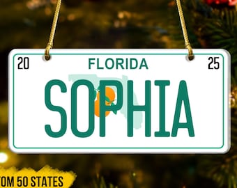 May include: A rectangular ornament resembling a Florida license plate, featuring the name "SOPHIA" in teal letters. The word "FLORIDA" is above the name, with the years "20" and "25" on either side. The ornament is suspended by a gold string.