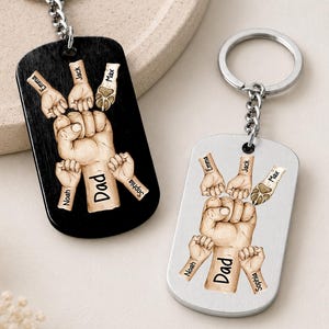 May include: Two keychains with a fist design, one black and one silver. The fist is surrounded by smaller hands and paw prints, with names like "Emma," "Jack," and "Max." The word "Dad" is printed on the fist. Each keychain has a silver ring.