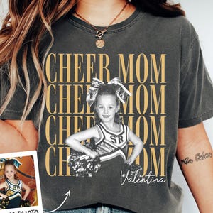 Puede incluir: Una camiseta gris carbón de manga corta con el texto dorado repetido CHEER MOM. Una fotografía en blanco y negro de una animadora con uniforme está centrada en la camiseta, con el nombre Valentina en letra cursiva blanca y una foto.
