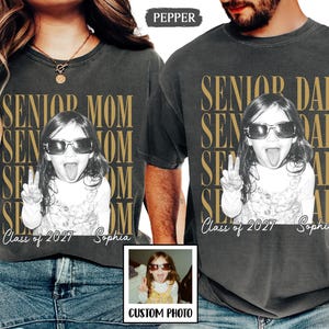 May include: Two charcoal gray t-shirts featuring a repeating gold text design that reads SENIOR MOM and SENIOR DAD. Each shirt displays a black and white photograph of a child wearing sunglasses and making a peace sign, with Class of 2027 and Sophia printed below.