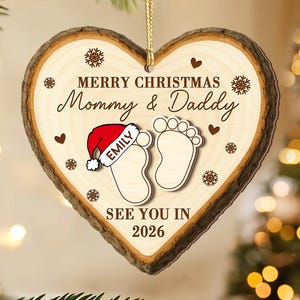 May include: Heart-shaped wooden Christmas ornament with a natural bark edge. The ornament features the text "MERRY CHRISTMAS Mommy & Daddy" and "SEE YOU IN 2026". It includes a Santa hat and footprints, with the name "EMILY".