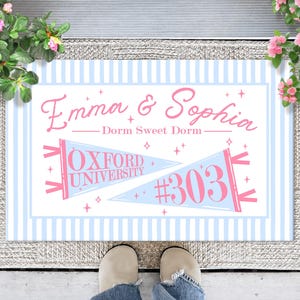 May include: A rectangular doormat with a light blue and white vertical striped border. The center features the pink text Emma & Sophia, the phrase Dorm Sweet Dorm, and two pennants. One pennant reads Oxford University, the other displays #303.