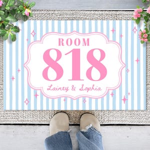 May include: A rectangular doormat featuring a light blue and white vertical striped pattern with a central white scalloped frame. The text ROOM 818 is printed in large pink letters, with the names Lainey & Sophia below in pink script.