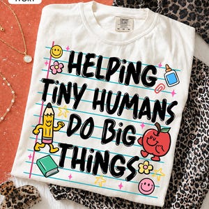 May include: An ivory t-shirt featuring a graphic of lined notebook paper with the text HELPING TINY HUMANS DO BIG THINGS in black. The design includes a yellow pencil, a red apple, a green book, a glue bottle, a paperclip, and smiley faces.