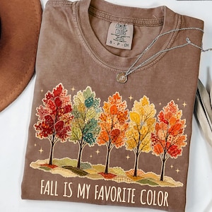 May include: An espresso brown t-shirt featuring a graphic of five trees with patterned autumn leaves in red, green, orange, and yellow. The text "FALL IS MY FAVORITE COLOR" is printed in white below the trees. A silver necklace rests on the shirt.