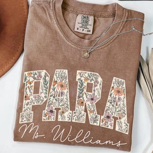 Puede incluir: Una camiseta color espresso con cuello redondo y etiqueta Comfort Colors. El frente presenta la palabra PARA en letras grandes de bloque rellenas con un patrón floral, y el texto Ms. Williams en letra cursiva blanca debajo.