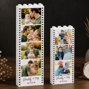 May include: Two white Lego-style photo blocks, one tall and one short, each displaying family photos. The blocks have a film strip design with the text "Daddy + Me" and the year "2026" in red. The photos show a father and baby.