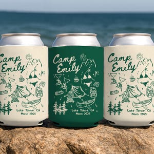 May include: Three can coolers with camping-themed designs. Two are off-white, and one is dark green. Each features the text "Camp Emily" in a script font, along with camping illustrations and the text "Lake Tahoe, CA March 2025."