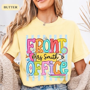May include: A butter yellow t-shirt featuring a colorful graphic with the text FRONT OFFICE and Mrs. Smith. The design includes a yellow pencil, a globe, a glue bottle, and small flowers against a background of light blue vertical stripes.