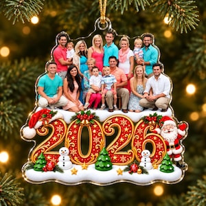 Puede incluir: Un adorno navideño de acrílico transparente con una foto grupal de dieciséis personas. Debajo de la foto, el año 2026 se muestra en grandes números rojos y dorados ornamentados, decorado con dos muñecos de nieve, tres árboles verdes y Santa Claus.