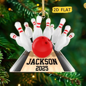 May include: A 2D flat bowling ornament featuring a red bowling ball and white pins with red stripes. The ornament has the text "JACKSON 2025" on the lane. The ornament is hanging from a gold string.
