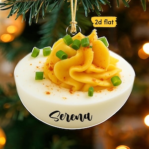 May include: A deviled egg ornament with a yellow filling, sprinkled with paprika and green chives. The ornament has the name "Serena" in a cursive font. The background is blurred with Christmas tree branches and lights.