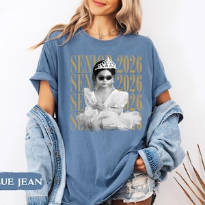 K&ouml;nnte beinhalten: Blaues Jeans-T-Shirt mit einem Schwarz-Wei&szlig;-Bild eines Kindes mit Sonnenbrille und Tiara. Der Text "SENIOR 2026" ist vertikal in Gold auf dem Shirt aufgedruckt.