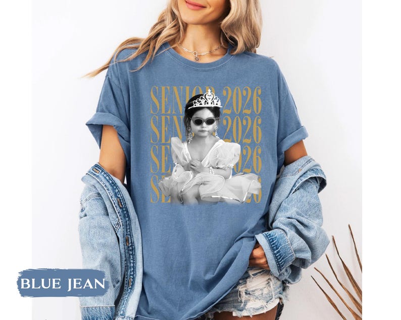 K&ouml;nnte beinhalten: Blaues Jeans-T-Shirt mit einem Schwarz-Wei&szlig;-Bild eines Kindes mit Sonnenbrille und Tiara. Der Text "SENIOR 2026" ist vertikal in Gold auf dem Shirt aufgedruckt.