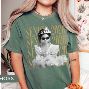 K&ouml;nnte beinhalten: Moosgr&uuml;nes T-Shirt mit einem Aufdruck eines Kindes in einem Tutu, Sonnenbrille und Diadem, mit dem Text "SENIOR 2026" in Gold. Das Shirt hat eine lockere Passform.