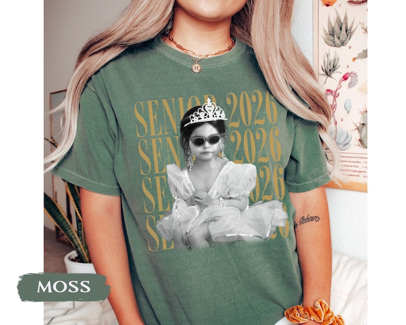 K&ouml;nnte beinhalten: Moosgr&uuml;nes T-Shirt mit einem Aufdruck eines Kindes in einem Tutu, Sonnenbrille und Diadem, mit dem Text "SENIOR 2026" in Gold. Das Shirt hat eine lockere Passform.