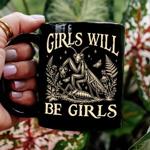 May include: A black ceramic mug featuring a cream-colored graphic of a praying mantis standing over another insect. The text "GIRLS WILL BE GIRLS" is printed in a decorative font above and below the illustration, surrounded by small botanical elements.