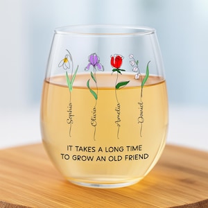 May include: A stemless wine glass filled with a light-colored liquid. The glass is decorated with line art flowers and names: Sophia, Olivia, Amelia, and Daniel. The text "IT TAKES A LONG TIME TO GROW AN OLD FRIEND" is printed on the glass.