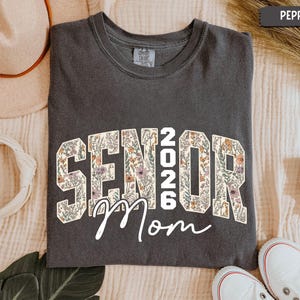 May include: A dark grey t-shirt featuring the text "SENIOR 2026 Mom" in a floral pattern. The t-shirt is a crew neck style. White trainers are visible at the bottom of the image.