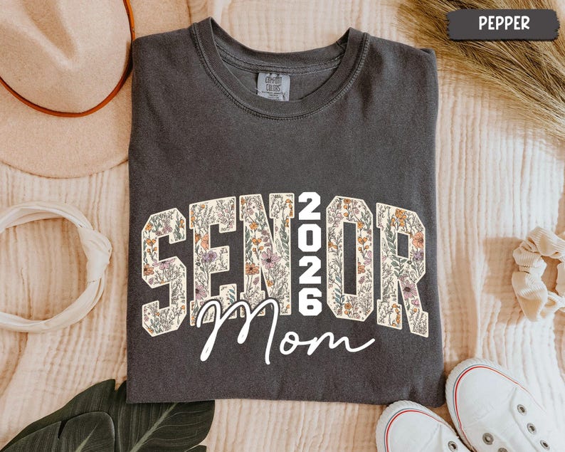 May include: A dark grey t-shirt featuring the text "SENIOR 2026 Mom" in a floral pattern. The t-shirt is a crew neck style. White trainers are visible at the bottom of the image.