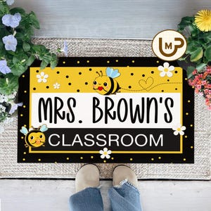 May include: A rectangular doormat with a black border and a yellow and black polka dot top section. The mat features the text "MRS. BROWN'S CLASSROOM" in large black letters, along with bee and flower graphics.