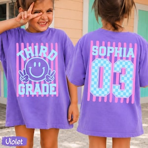 May include: A violet t-shirt featuring a front graphic with pink vertical stripes, a light blue smiley face, and the text "THIRD GRADE". The back displays the name "SOPHIA" and the number "03" in light blue with a checkered pattern.