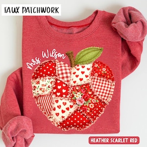 May include: Heather scarlet red sweatshirt with a faux patchwork apple design. The apple features various red patterns, including polka dots, hearts, and floral prints. The text "Mrs. Wilson" is written on the sweatshirt.