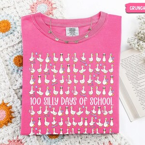K&ouml;nnte beinhalten: Ein pinkfarbenes Comfort Colors T-Shirt mit einem Muster aus Cartoon-G&auml;nsen mit Sonnenbrillen, Luftballons und Skateboards. Der Text "100 Silly Days of School" ist darunter aufgedruckt. Eine silberne Halskette ist &uuml;ber dem T-Shirt drapiert.