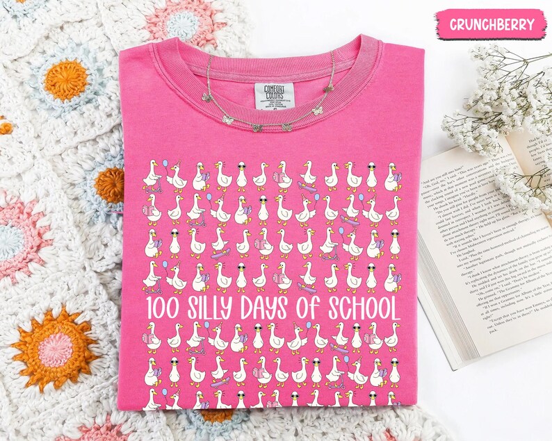 K&ouml;nnte beinhalten: Ein pinkfarbenes Comfort Colors T-Shirt mit einem Muster aus Cartoon-G&auml;nsen mit Sonnenbrillen, Luftballons und Skateboards. Der Text "100 Silly Days of School" ist darunter aufgedruckt. Eine silberne Halskette ist &uuml;ber dem T-Shirt drapiert.