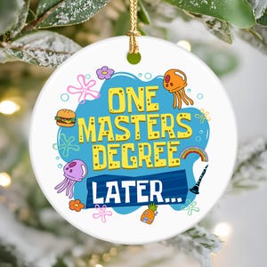 May include: A white ceramic ornament with a gold string for hanging. The ornament features the text "ONE MASTERS DEGREE LATER..." in yellow, surrounded by cartoon illustrations of a burger, jellyfish, and a pineapple house.