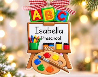 May include: A hanging ornament shaped like an easel with a white board displaying the text Isabella Preschool and 2026. It features colourful ABC blocks, a crayon cup, paint pots, and a wooden palette with a paintbrush, topped with a red gingham bow.