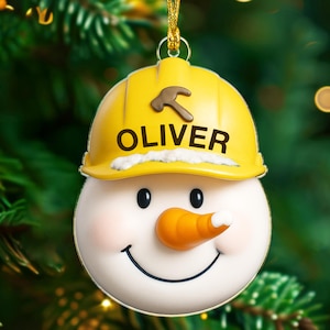 May include: A snowman Christmas ornament with a yellow construction hat featuring a brown hammer and the name "OLIVER". The snowman has a carrot nose, black eyes, and a smiling mouth. The ornament hangs from a gold string.