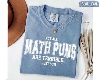 May include: A blue jean colored t-shirt with white text that reads NOT ALL MATH PUNS ARE TERRIBLE... JUST SUM. The shirt features a crew neckline and a Comfort Colors brand label on the inside collar. A gold-toned necklace rests on the fabric.